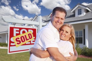 Happy Couple Hugging in Front of Sold Real Estate Sign and House.